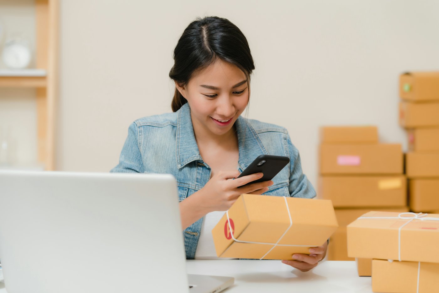 A store owner packing an order while checking her phone, with boxes and a laptop on the table
